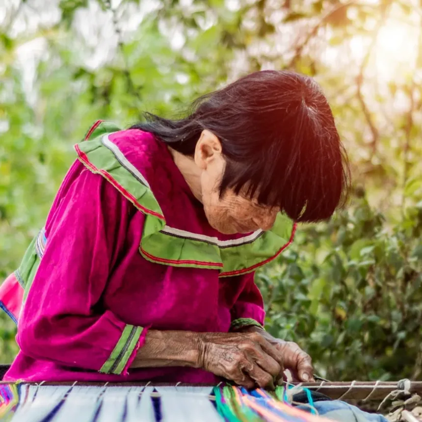 An elderly woman in a magenta blouse with green trim bends over her loom, her hands working brightly coloured threads, outdoors under trees